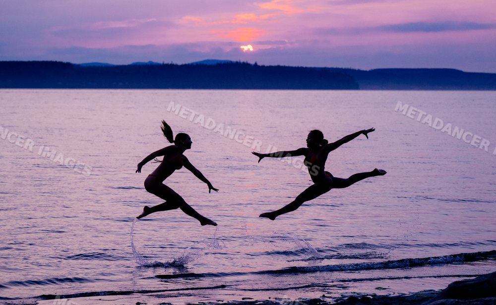 Two women perform acrobatic jumps in water at sunset on lake shore