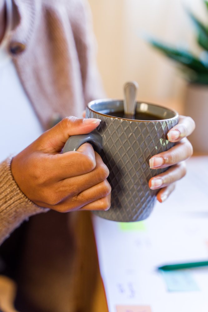 Hands holding a gray textured mug containing coffee with a spoon inside near a table with paper and pen indoors, Spain.
