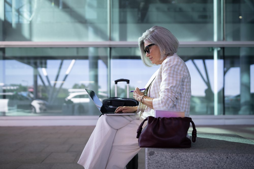 Mature businesswoman with gray hair and sunglasses working on laptop and checking notes in airport seating area beside luggage and handbag, Spain, Madrid.
