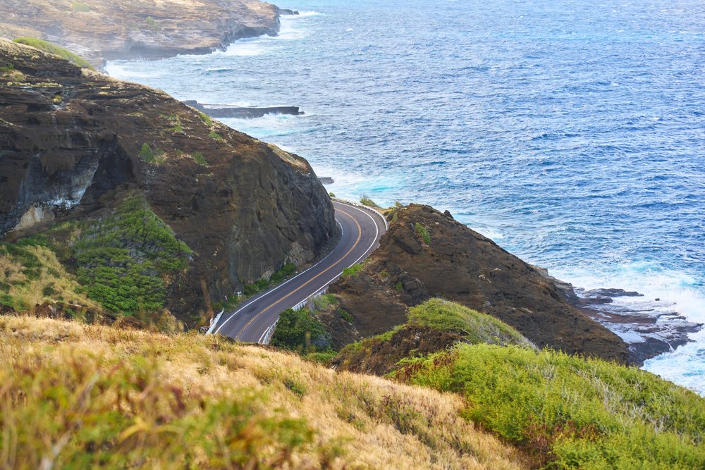 Scenic coastal road winding around rocky cliffs with ocean waves and vegetation, Oahu, Hawaii, USA.