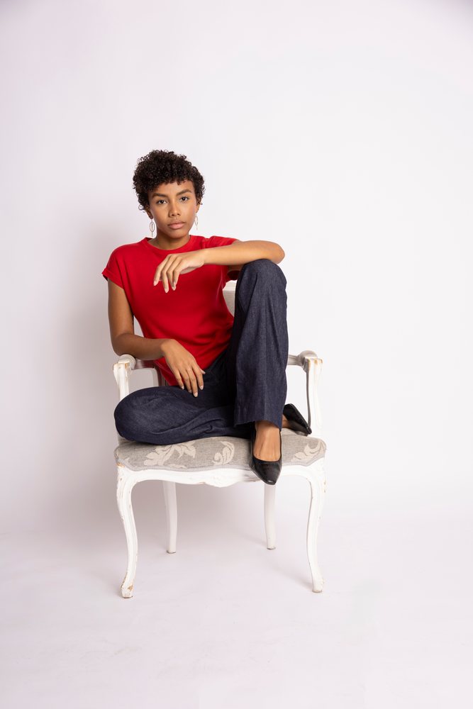 Young woman with curly hair and earrings sits on a white chair wearing a red shirt, blue pants, and black shoes in a studio setting, fashion, young culture.