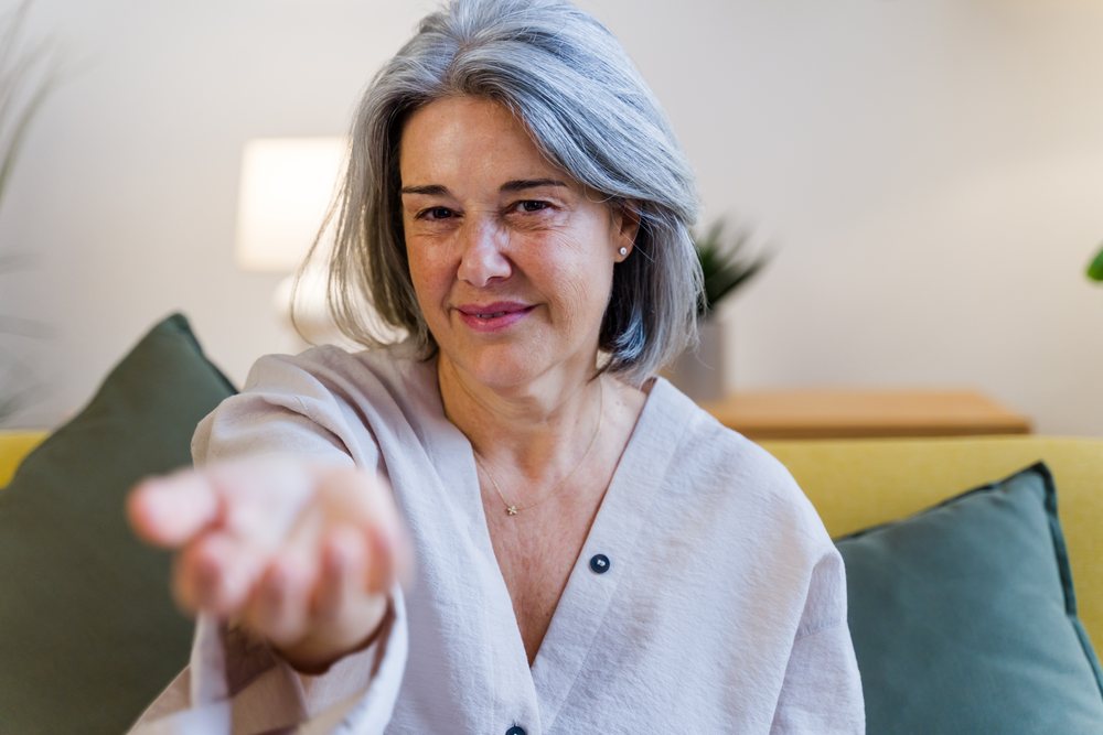 Middle-aged woman with gray hair sitting on a sofa indoors, reaching out her hand with a smile, Spain.