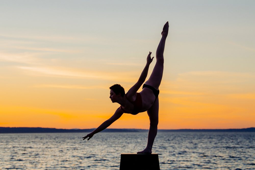 Woman performs a yoga balance pose on a platform by calm waters at sunset, USA.