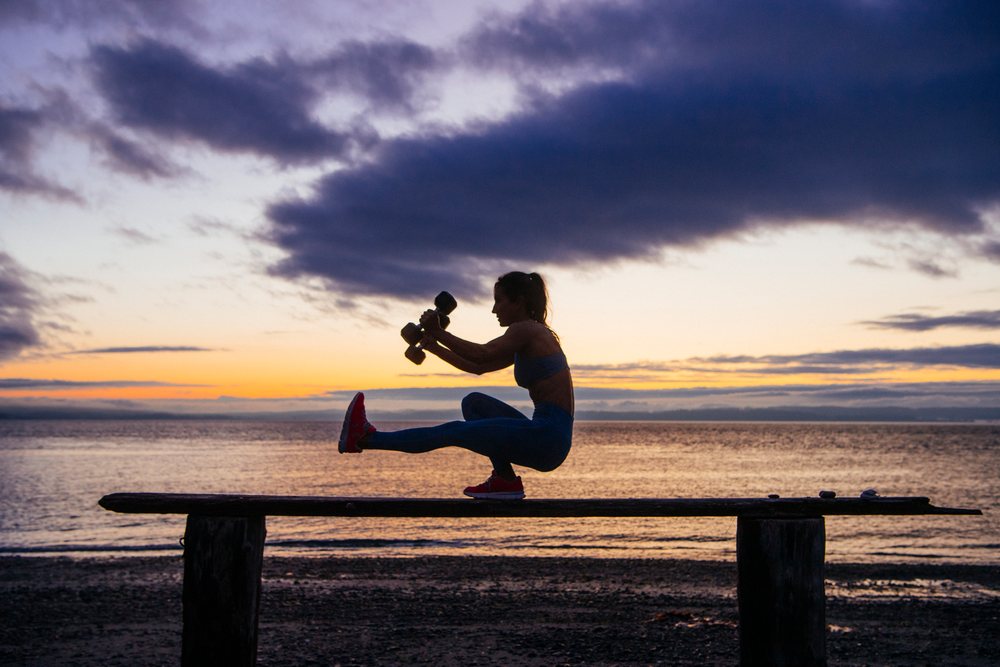 Woman holding dumbbells and balancing on a bench during an outdoor exercise at sunset on a beach, WA, USA.
