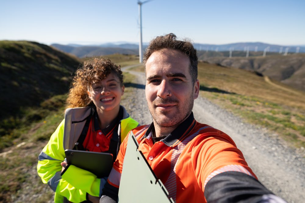 Two people wearing safety jackets take a selfie on a trail near wind turbines amid rolling hills, sustainability, renewable energy, Galicia, Spain.