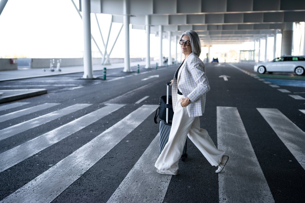Mature woman wearing glasses pulls a rolling suitcase with a black bag on top while walking on a pedestrian crossing in urban, businesswoman, Spain, Madrid.