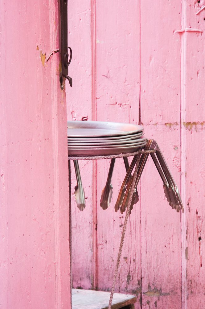 Metal trays and tongs hanging on a holder behind a rustic pink wooden door with peeling paint in a kitchen setting, Guanajuato, Mexico.