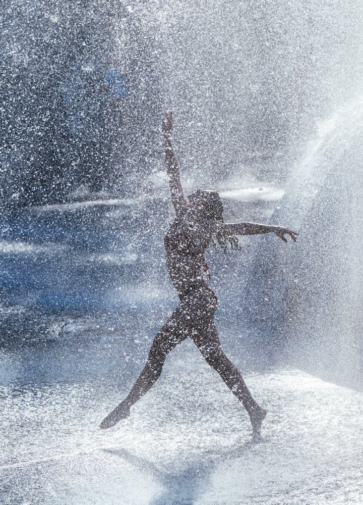Female dancer jumping gracefully through water spray in an outdoor setting in the USA with a theater advertisement banner in the background, partner, acrobatics, yoga.