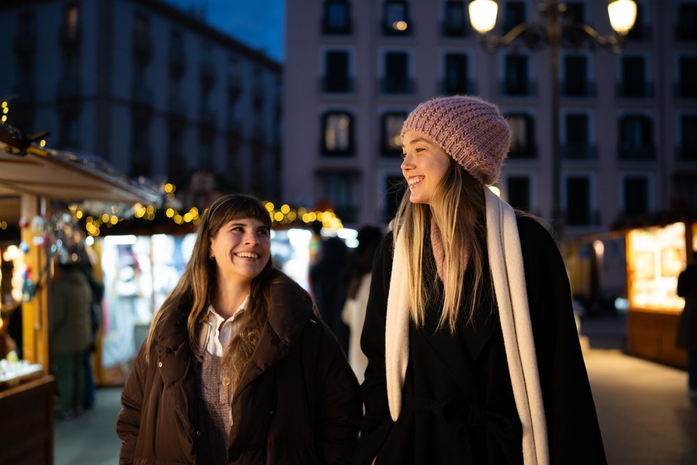 Two women smiling and walking together at a Christmas market in Madrid at night, Spain.