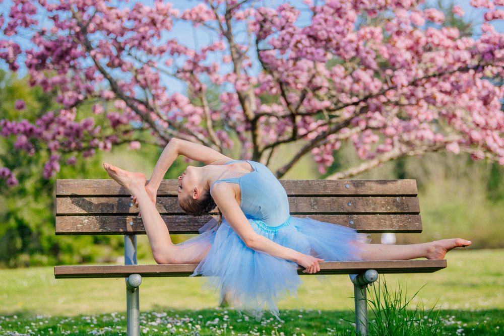 Female ballet dancer in blue tutu stretches back on a wooden bench amid blooming cherry blossoms in a USA park during spring.