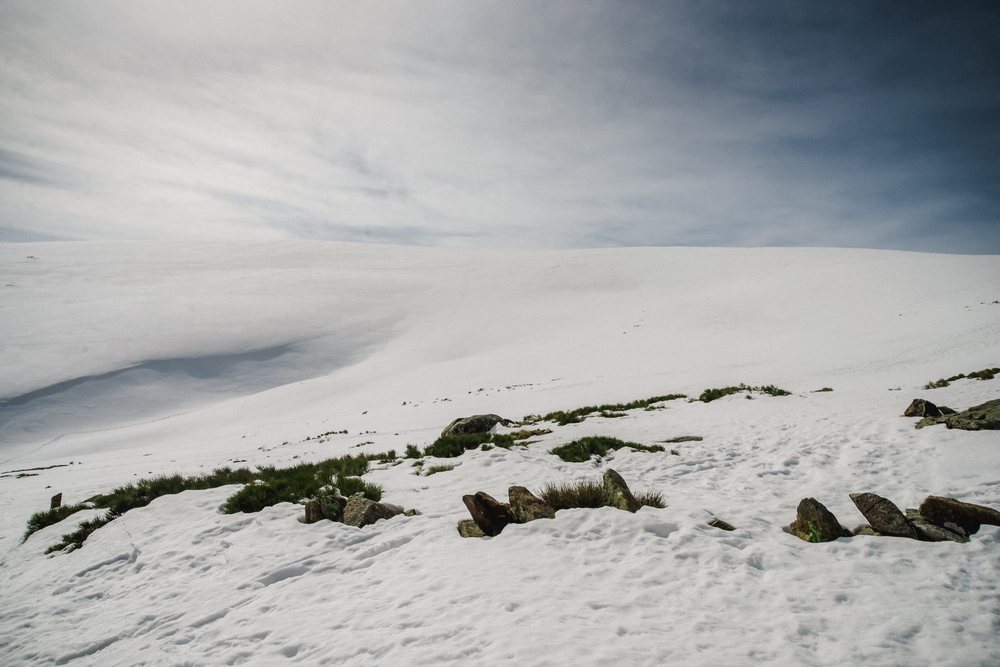 Snow-covered mountain terrain with scattered rocks and grass patches under a cloudy sky, mountaineering, climbing, landscape, Spain.