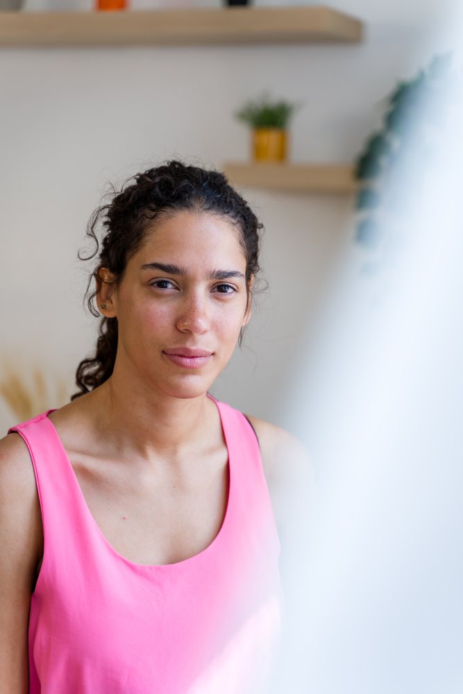 A young woman with curly hair wearing a pink sleeveless top is photographed indoors with a calm and confident expression in a home setting, Spain.