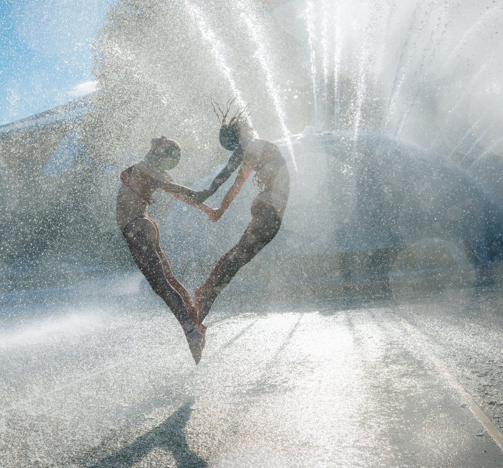 Two women jumping and holding hands in an acrobatic pose inside a water fountain spray on a sunny day, partner, acrobatics, yoga, USA.