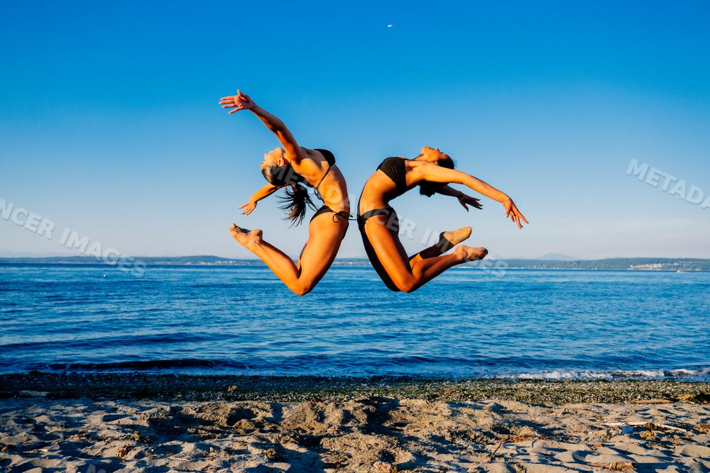 Two women jump in synchronized acrobatic motion on a beach in wa USA