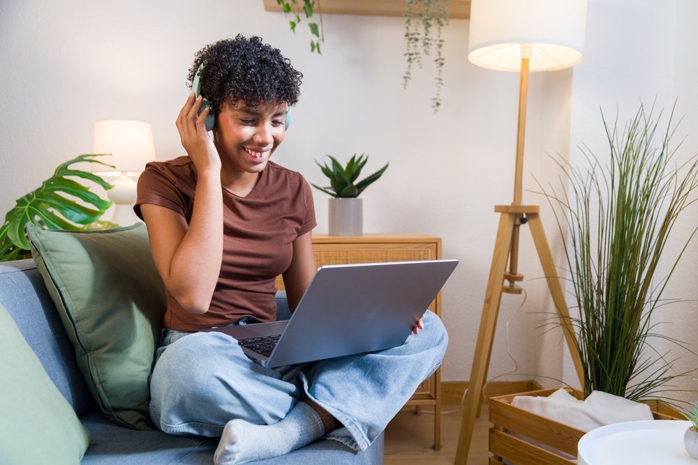 Young woman smiling and using a laptop while wearing headphones in a cozy home setting with plants and lamps.