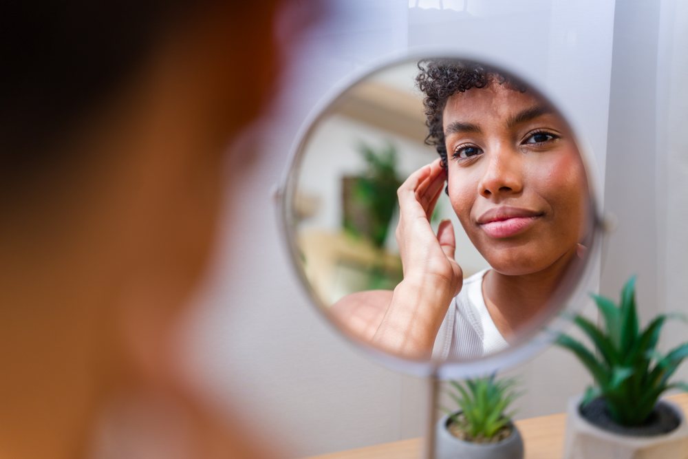 Woman with curly hair touches her face while looking into a round mirror on a table decorated with small indoor plants, Spain.