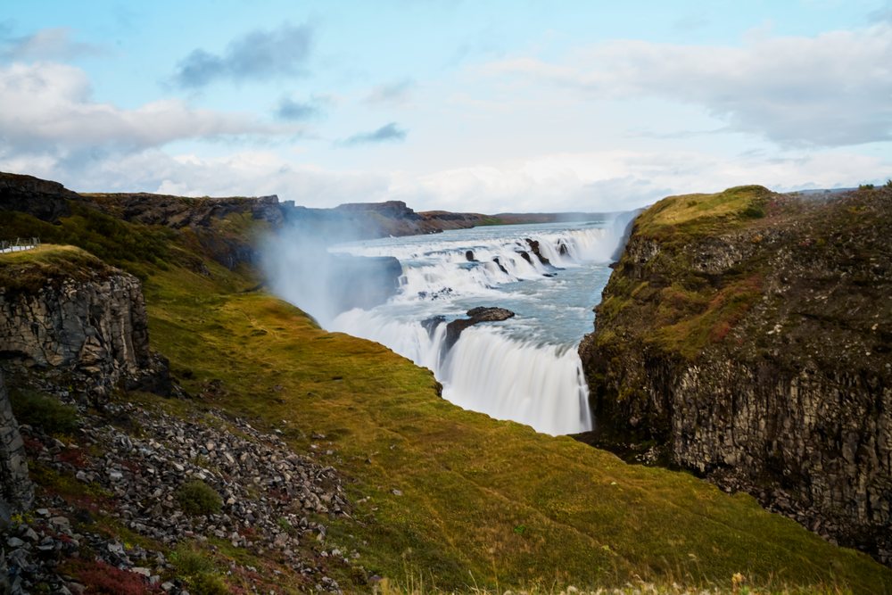 A powerful waterfall cascades between rugged basalt cliffs surrounded by green grass and rocky terrain, Iceland.