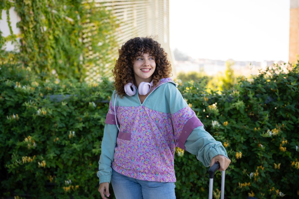 Young woman with curly hair wearing a floral hoodie and headphones smiles outdoors holding a suitcase handle in a green setting, Spain.