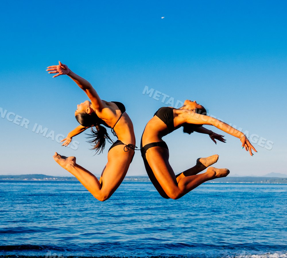 Two women in bikinis leap in synchronized backbend poses on a beach