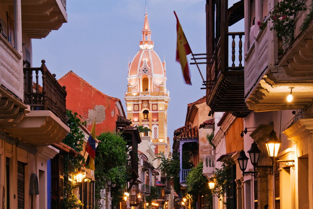 Cartagena cathedral tower rises above a historic street with colonial buildings and a Spanish flag at dusk, Colombia.