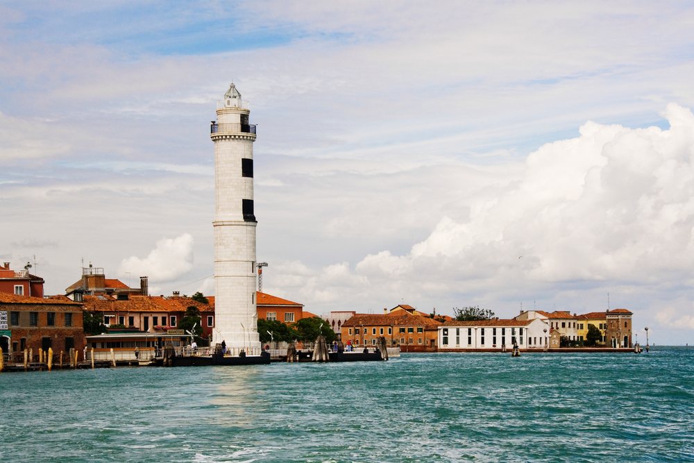 White lighthouse standing on a pier extending into the calm sea with red and white buildings near the shoreline and a cloudy sky in Italy.