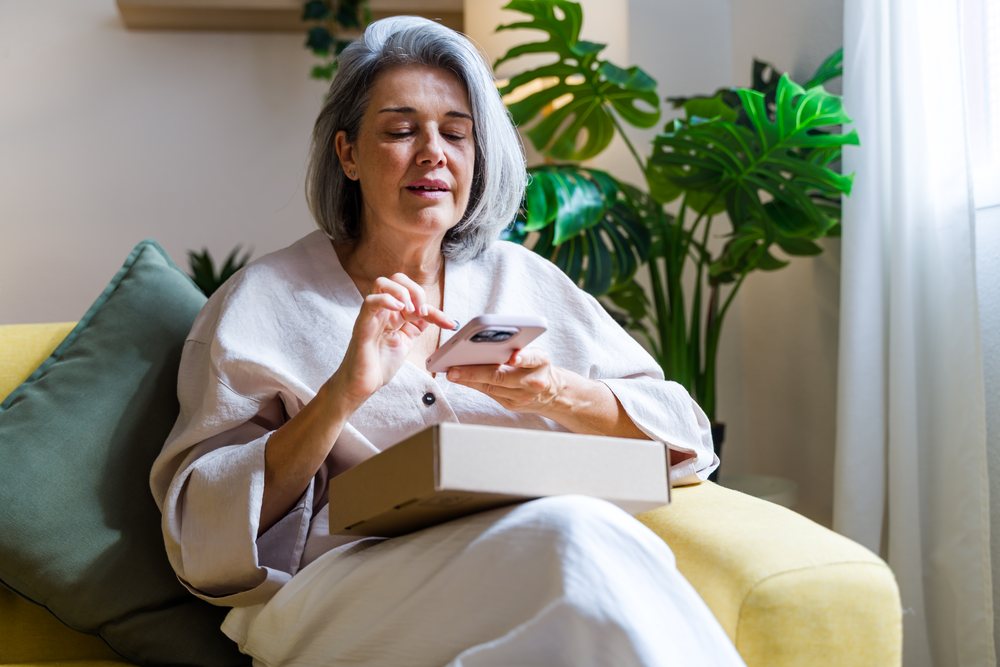 Middle-aged woman with gray hair using a smartphone while sitting on a yellow sofa with green cushions and indoor plants including monstera in a cozy home setting, Spain.