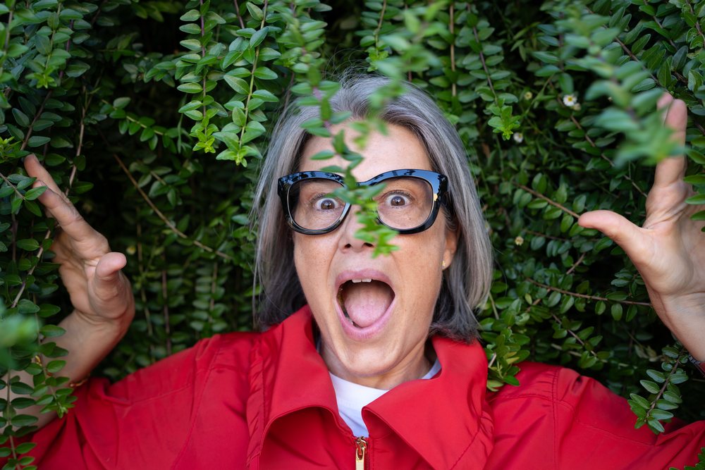 Woman with gray hair and glasses expressing surprise while holding green leaves outdoors, Spain.