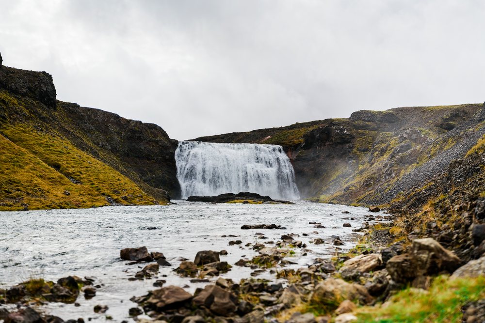 A waterfall flows into a rocky river surrounded by hillside terrain in Iceland under an overcast sky.