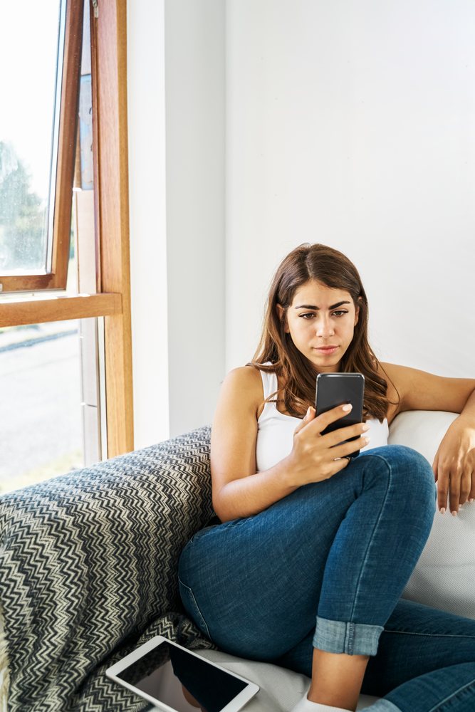 A white woman sits on a couch looking at her phone indoors