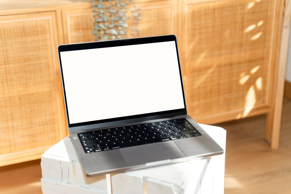 Laptop with a blank white screen on a white wooden desk in a sunlit interior with wooden furniture in the background, mock up, Spain.