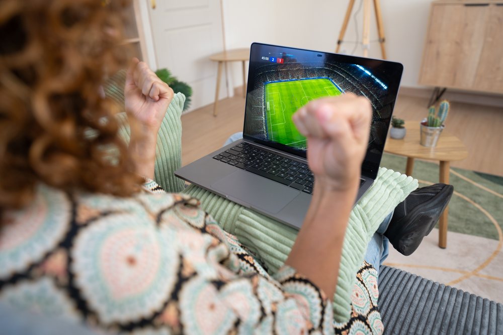 Woman with curly hair cheerfully watches a soccer match on a laptop while seated on a couch in her home, Spain.
