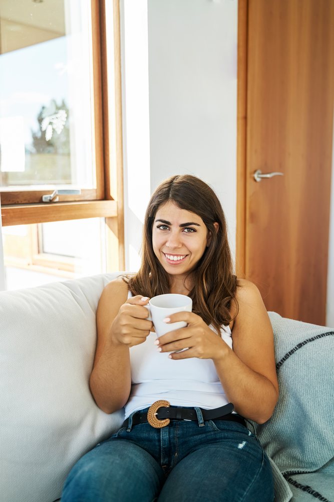 A young white woman sits on a couch holding a cup and smiling