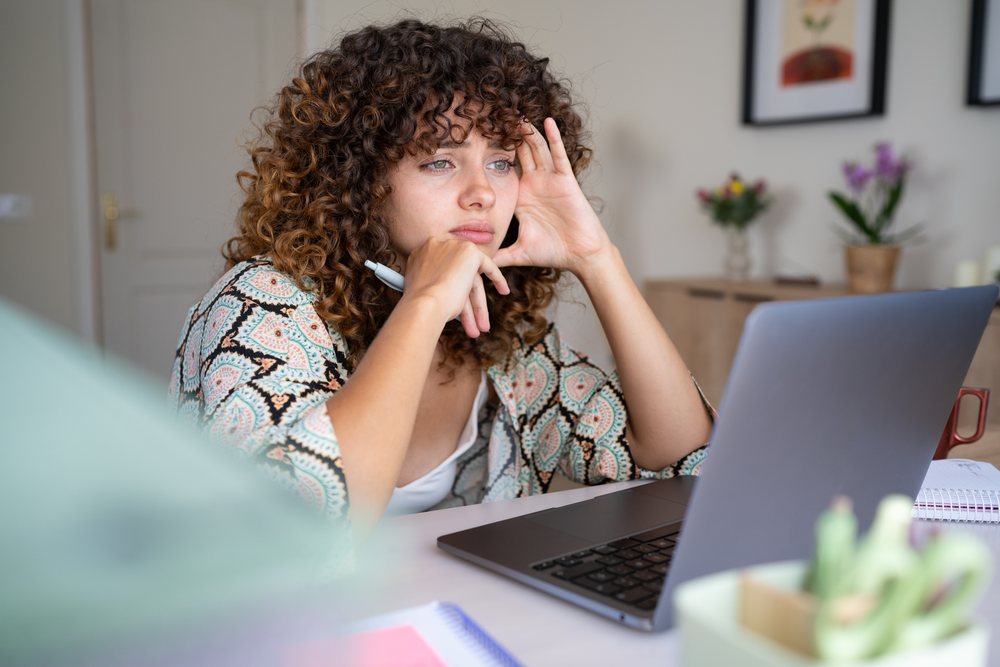 Girl with curly hair pensively looking at laptop screen while sitting at a desk in a home interior, Spain.