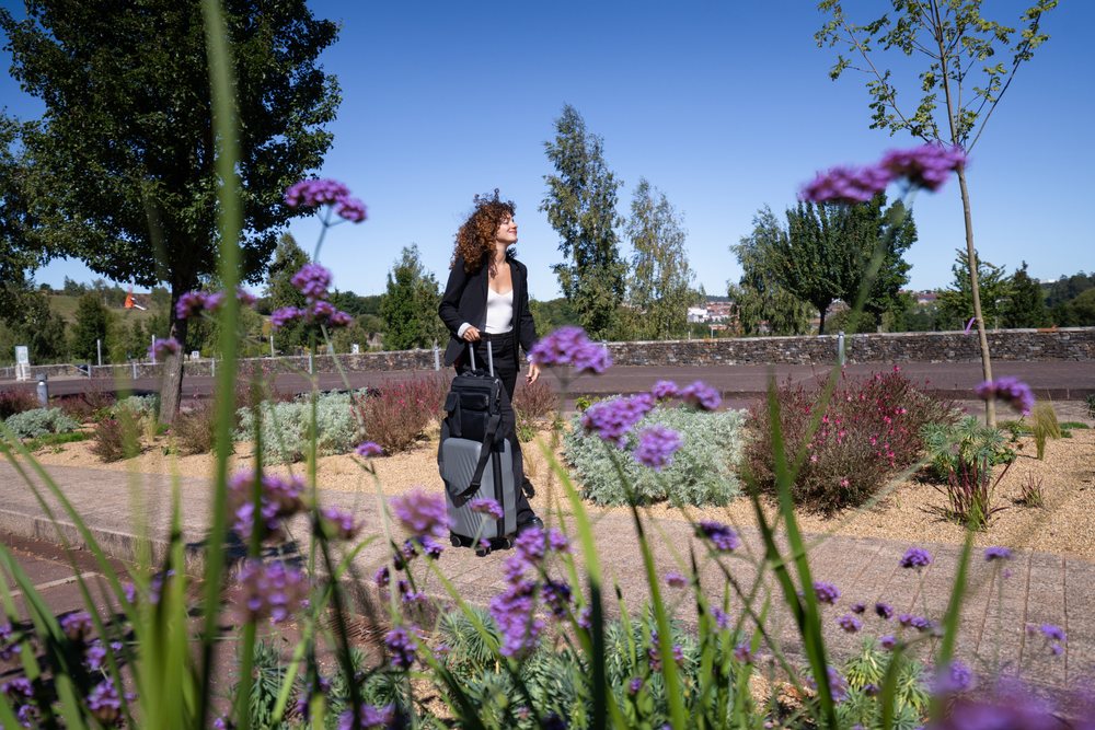 Woman walking outdoors in an urban park with flowering plants and rolling suitcase on a sunny day, Business Travel, Businesswoman, Luggage, Spain.