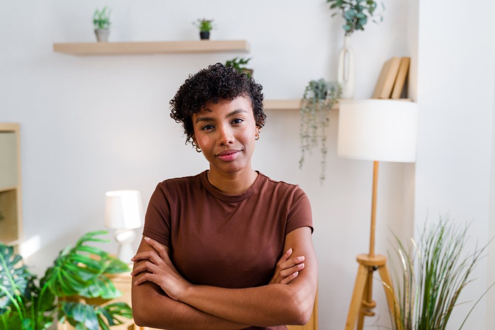 Confident woman with curly hair and brown shirt stands with crossed arms in a modern living room with plants and a floor lamp, Spain.