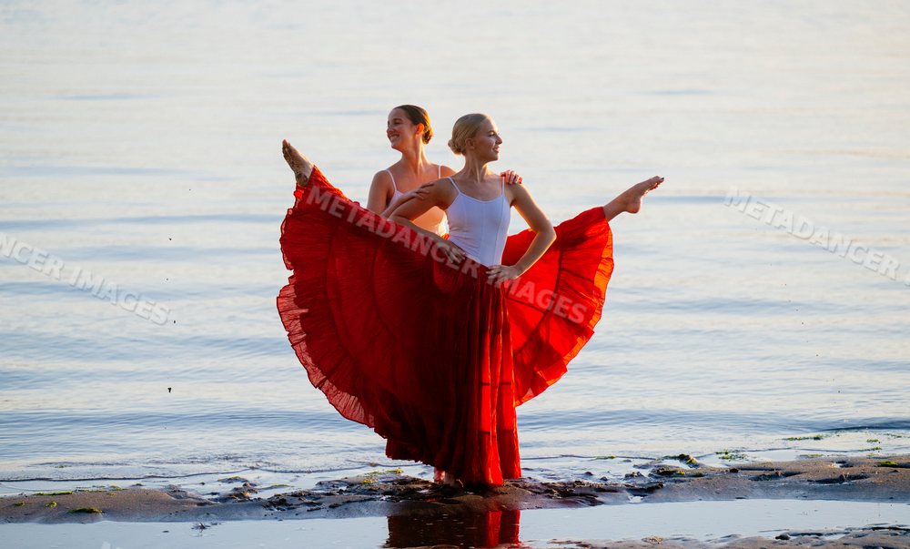 Two white women practice partner acrobatics on a beach at sunset