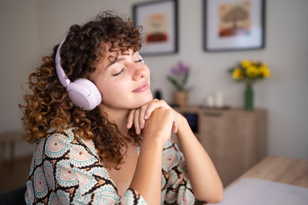 Woman with curly hair listens to music with headphones seated indoors at a table with a vase of flowers and decorated wall, home, girl, Spain.