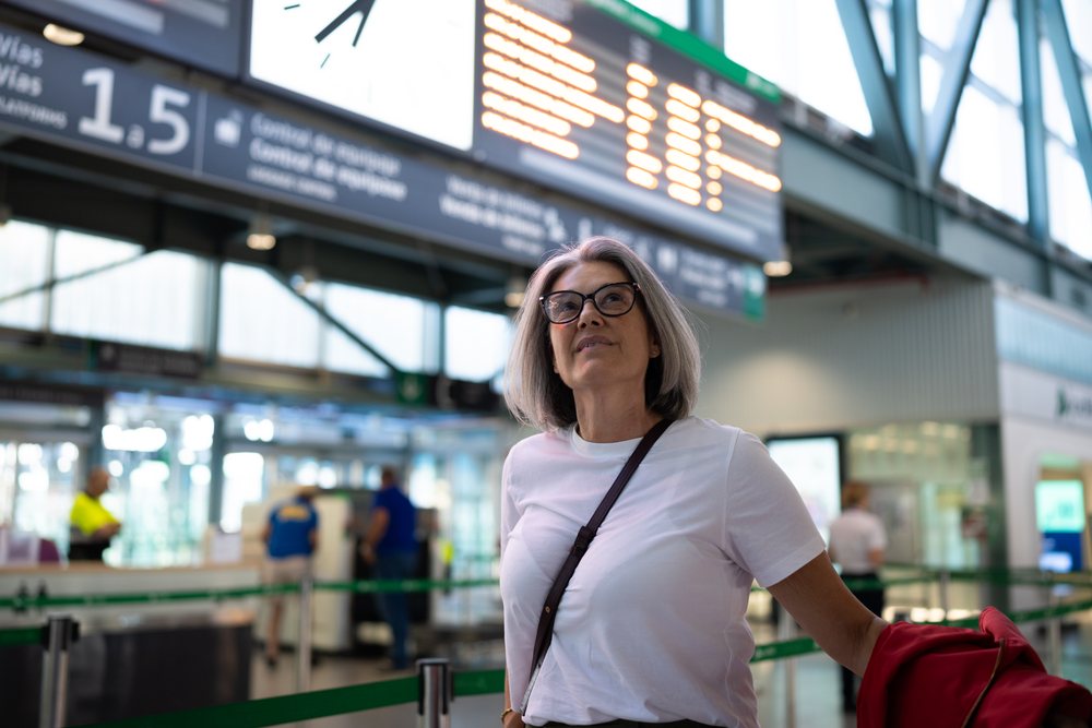 Woman with gray hair wearing glasses and white t-shirt holding a red jacket waits at the security checkpoint under a departure board inside a Spain airport terminal.