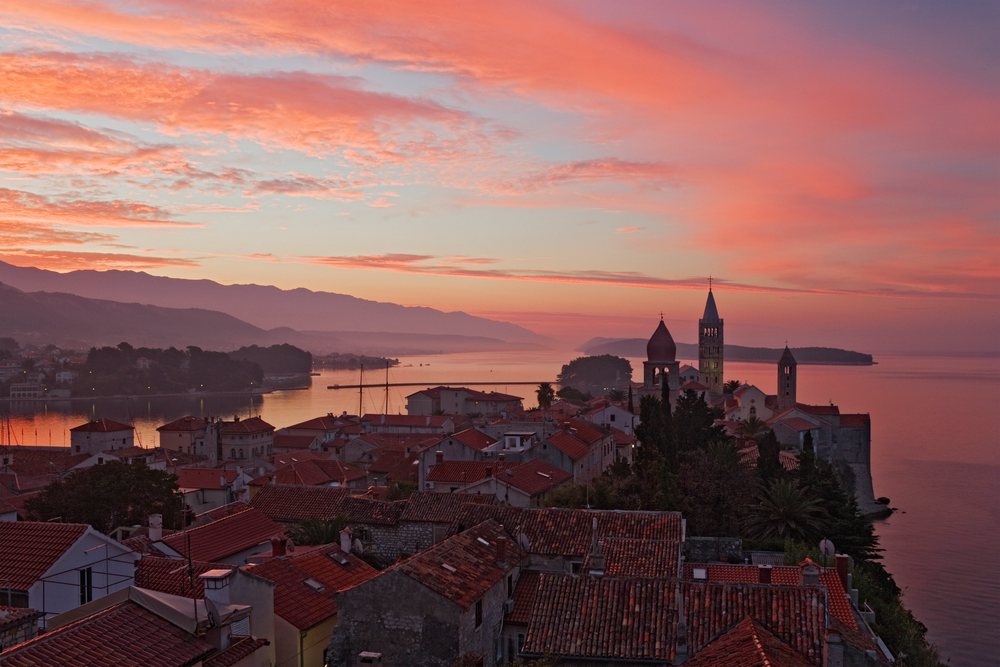 Dawn over Rab Island reveals a seaside town with red tile rooftops and bell towers beside calm water under a colorful sky, Croatia.
