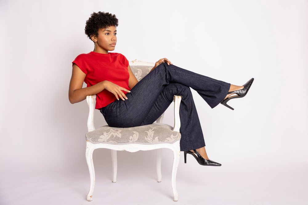 Woman with curly hair wearing a red top and black pants sitting on an ornate chair in a studio setting with a white background, fashion, clothes, young culture.