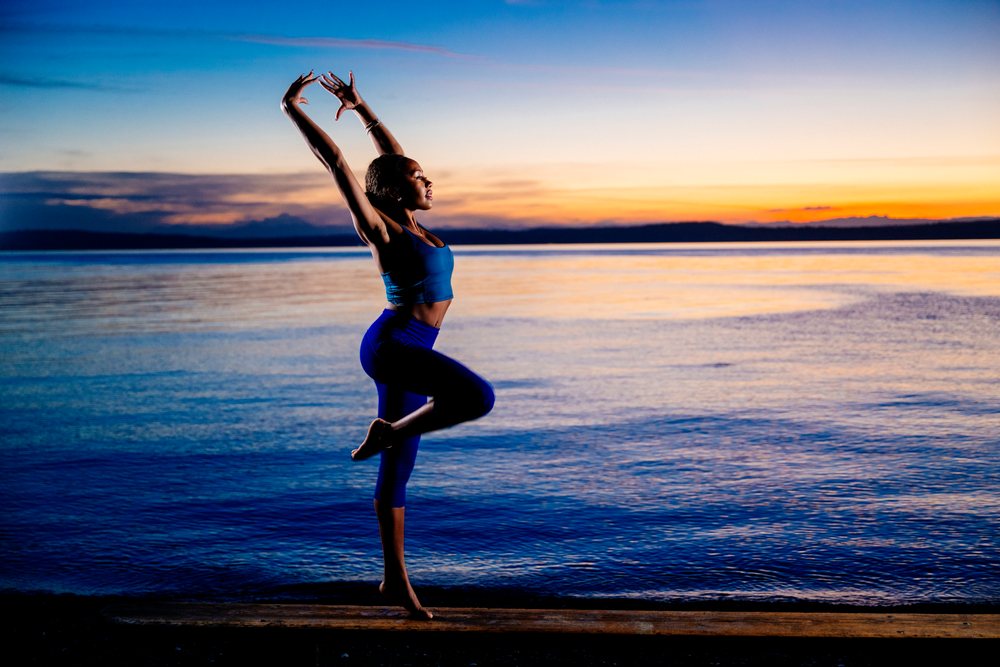 Woman performing a yoga balancing pose on one leg at the shore during a colorful sunset, USA.
