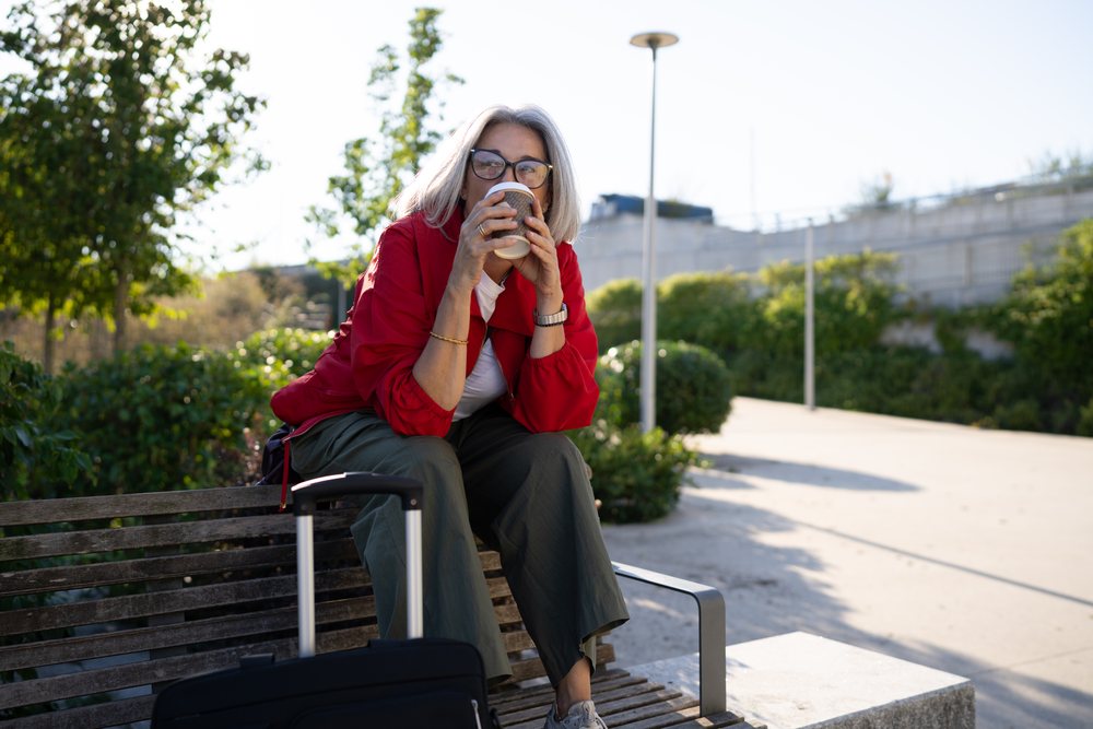 Woman with gray hair and glasses sits on a wooden bench in a park drinking coffee beside black wheeled luggage during daytime, Spain.