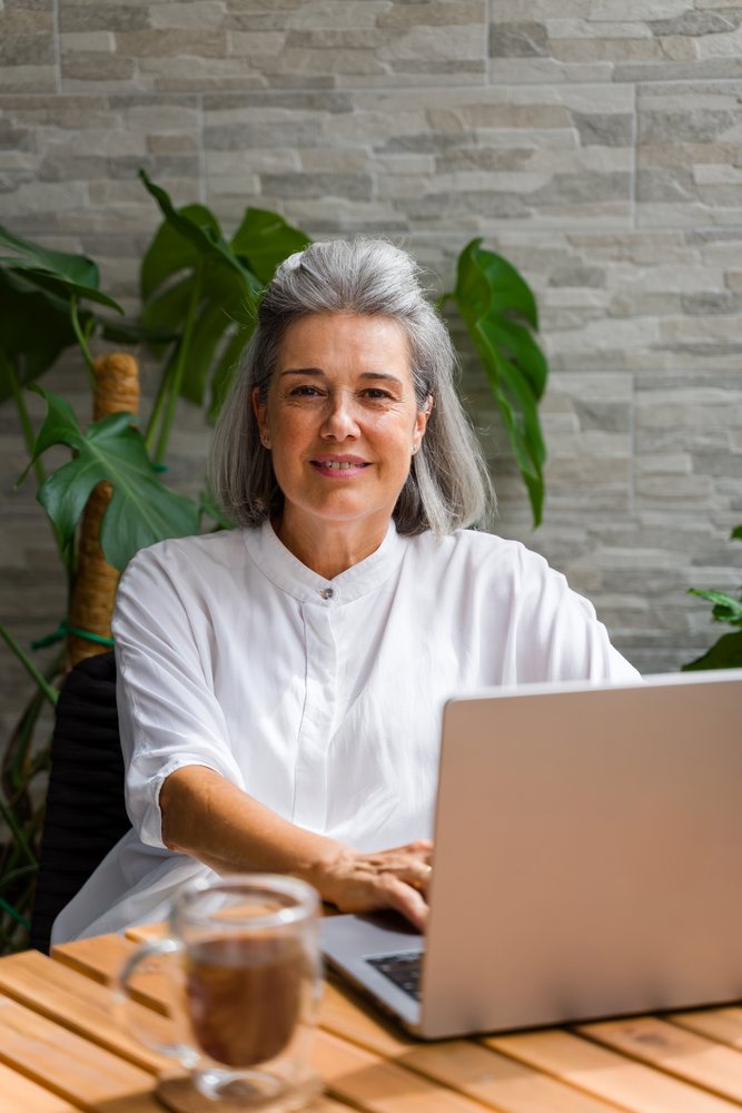 Woman working on a laptop with a cup of coffee at a wooden table indoors with plants in the background, Spain.