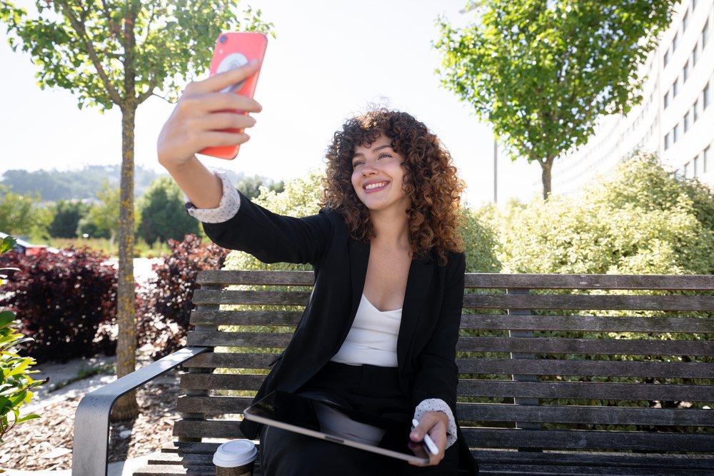 Woman sitting on an outdoor bench in Spain taking a selfie with her smartphone while holding a tablet, with a coffee cup nearby.