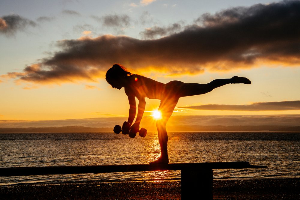Woman performing a balancing yoga pose holding dumbbells on a beach at sunset with calm water and scenic sky, WA, USA.