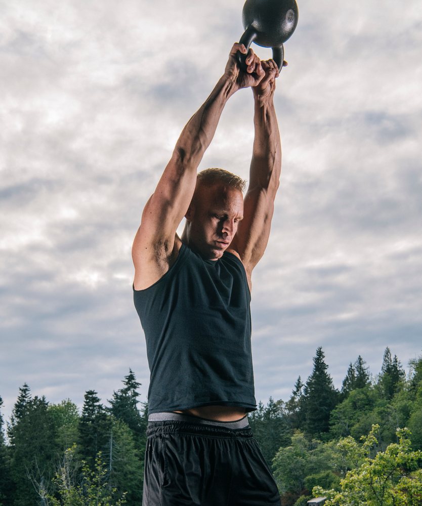 Man lifting a kettlebell overhead during an outdoor workout with trees and cloudy sky, USA.