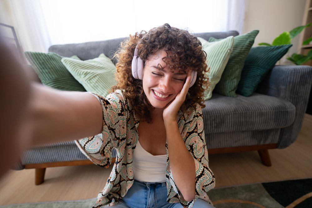 Woman with curly hair and headphones sitting on the floor smiling while taking a selfie in a living room with green pillows on the couch, home, girl, Spain.