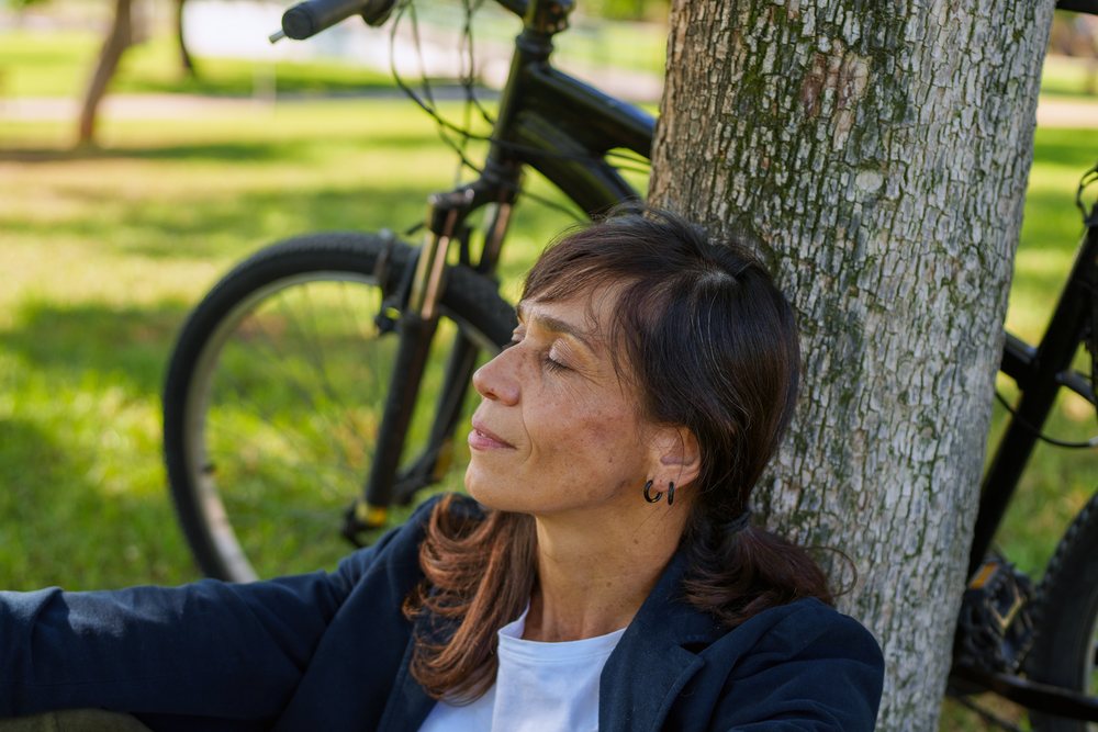 Middle-aged woman with dark brown hair wearing a dark jacket and white shirt resting against a tree near a black-framed bicycle in a sunny park, Málaga.