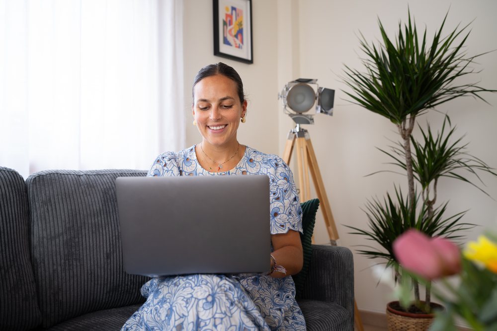 Woman in a blue and white floral dress smiles while working on a laptop seated on a gray sofa indoors, telecommuting, Spain.