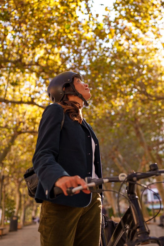Woman wearing a helmet stands beside a bicycle on a tree-lined park path in Málaga during autumn daylight.